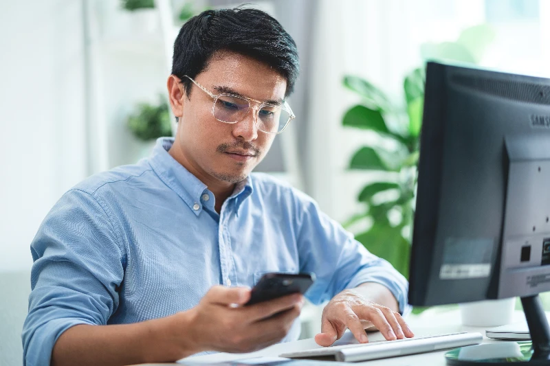 A man wearing glasses is sitting at a desk with a computer monitor and cell phone